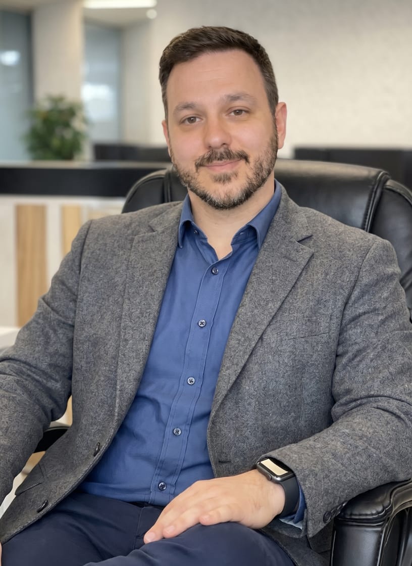 Jacob A man with short dark hair and a beard, wearing a grey blazer and blue shirt, sits in a black office chair, smiling slightly—an approachable face behind Insure My Health. The background shows a modern office with a plant and blurred furniture.